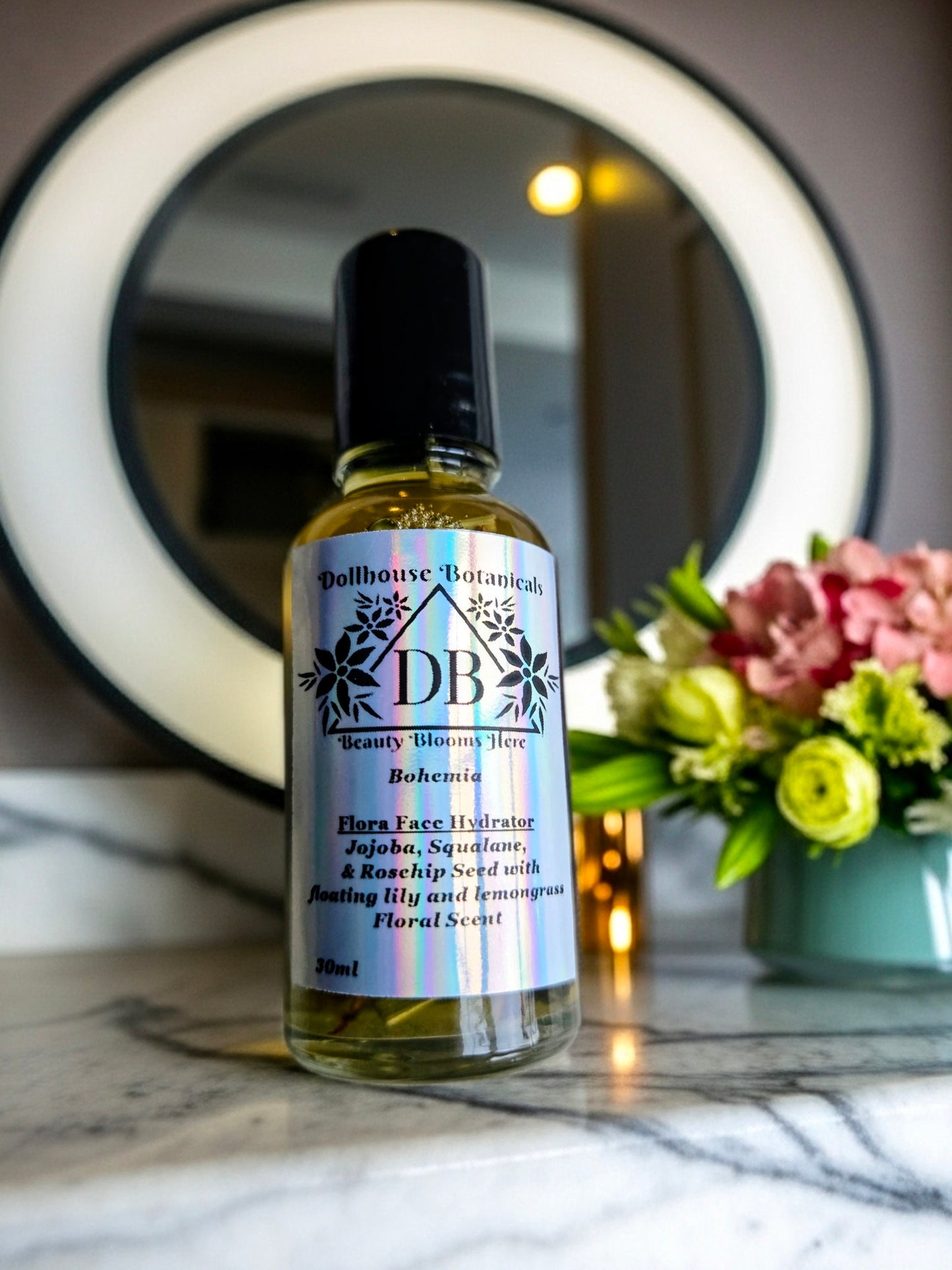 Bottle of Dollhouse Botanicals Bohemia Flora Face Hydrator. Golden oil with floating lemongrass and orange lily in glass bottle with steel roller ball applicator.
