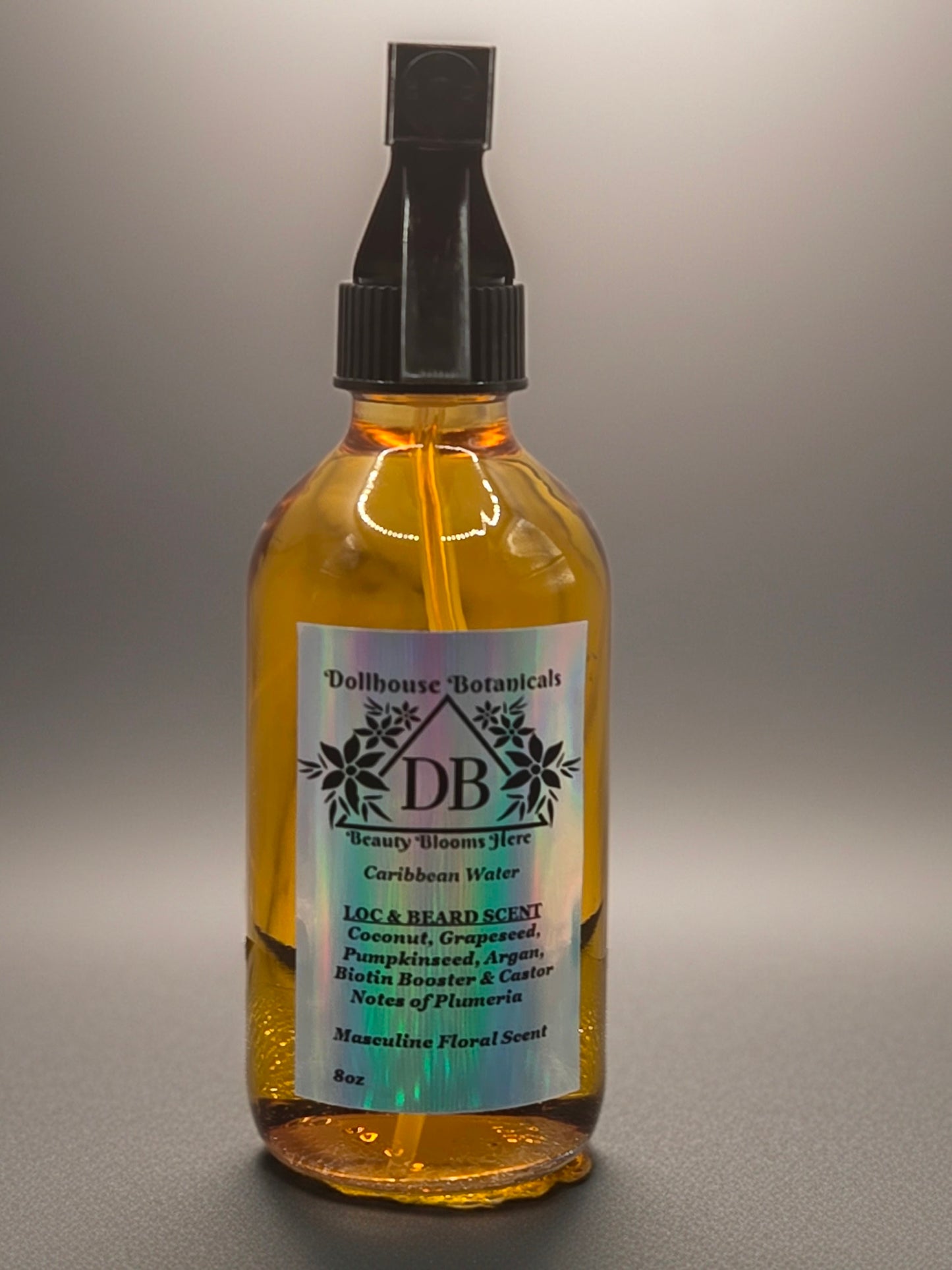 Bottle of Dollhouse Botanicals Caribbean Water Loc and Beard Scent. Golden oil in glass bottle with black trigger sprayer. Font facing with label and ingredients displayed.