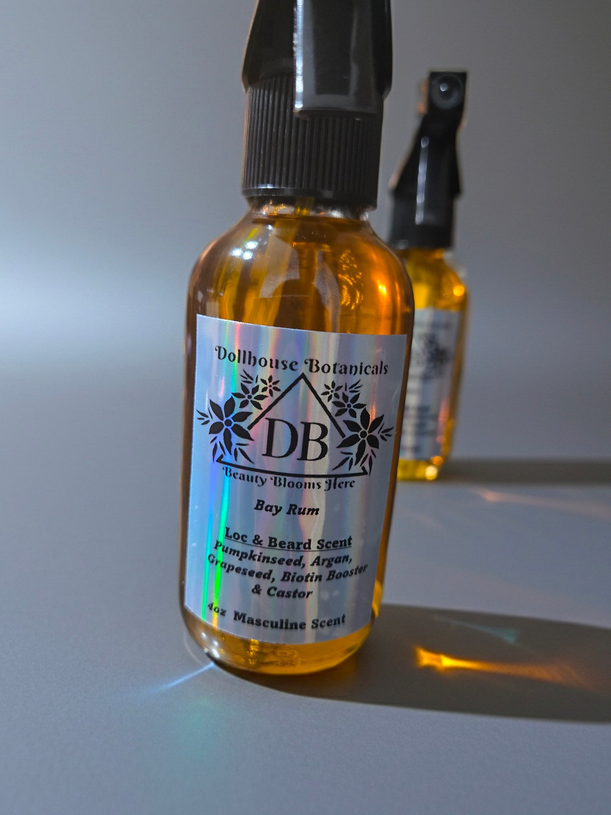 Bottle of Dollhouse Botanicals Bay Rum Loc and Beard Scent. Golden oil in glass bottle with trigger sprayer. Front facing with label and ingredients displayed.