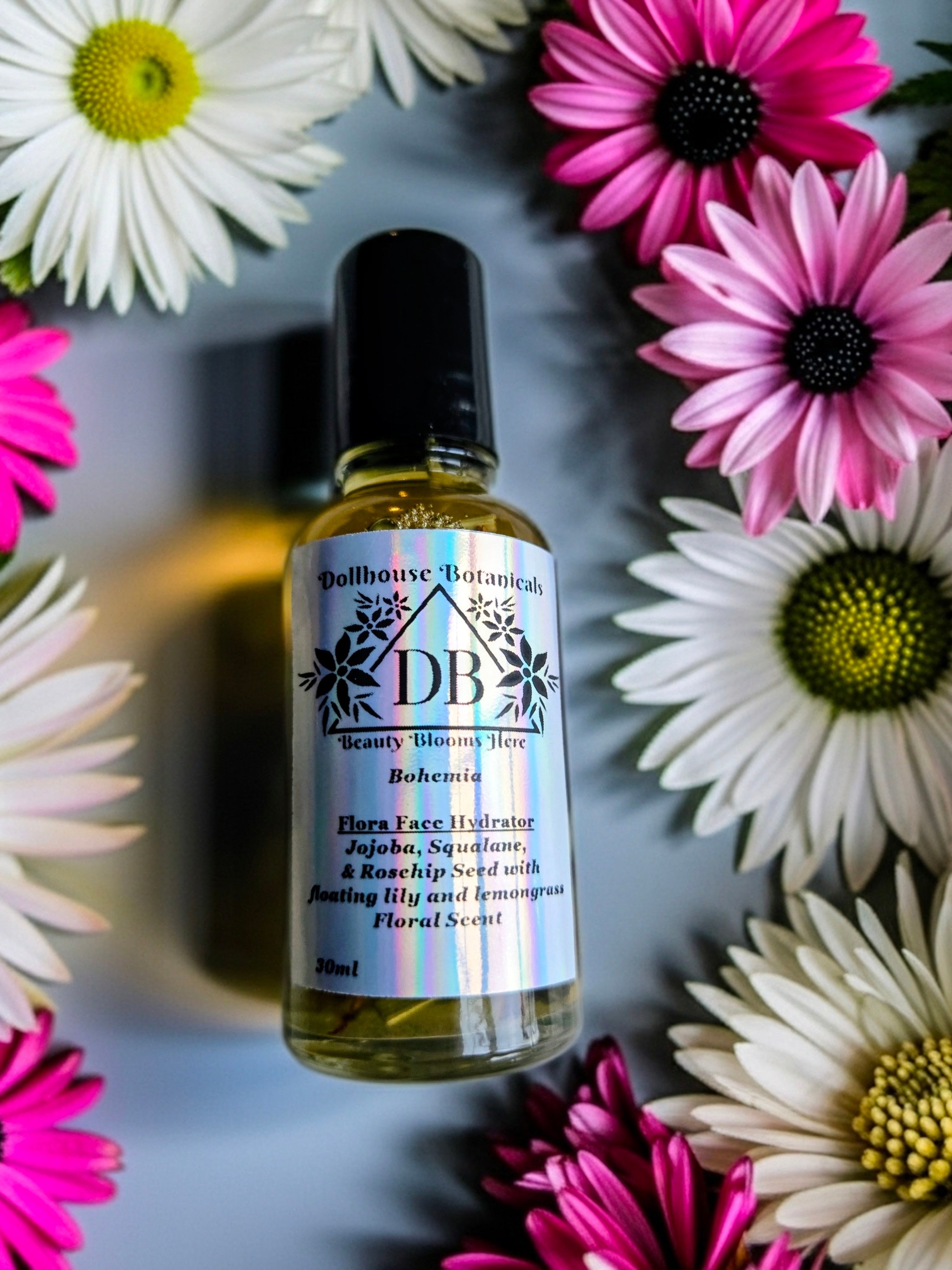 Bottle of Dollhouse Botanicals Bohemia Flora ace Hydrator with black top, lying on surface with flowers.