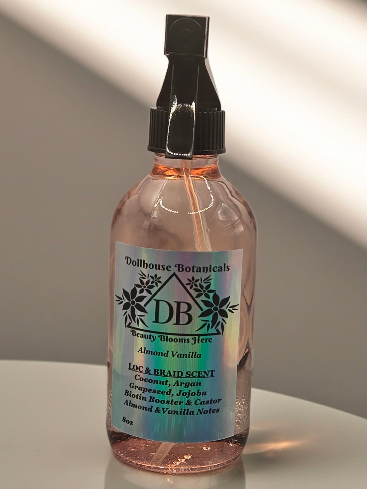 Bottle of Dollhouse Botanicals Almond Vanilla Loc and Braid Scent. Oil in glass bottle with trigger sprayer, sitting on pink surface. Front facing with label and ingredients displayed.