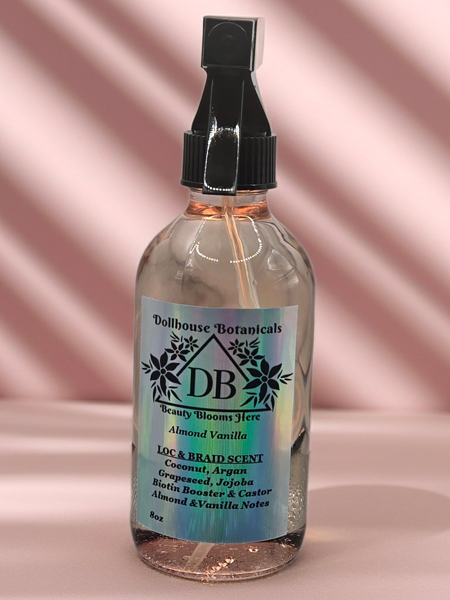 Bottle of Dollhouse Botanicals Almond Vanilla Loc and Braid Scent. Oil in glass bottle with trigger sprayer, sitting on pink surface. Front facing with label and ingredients displayed.