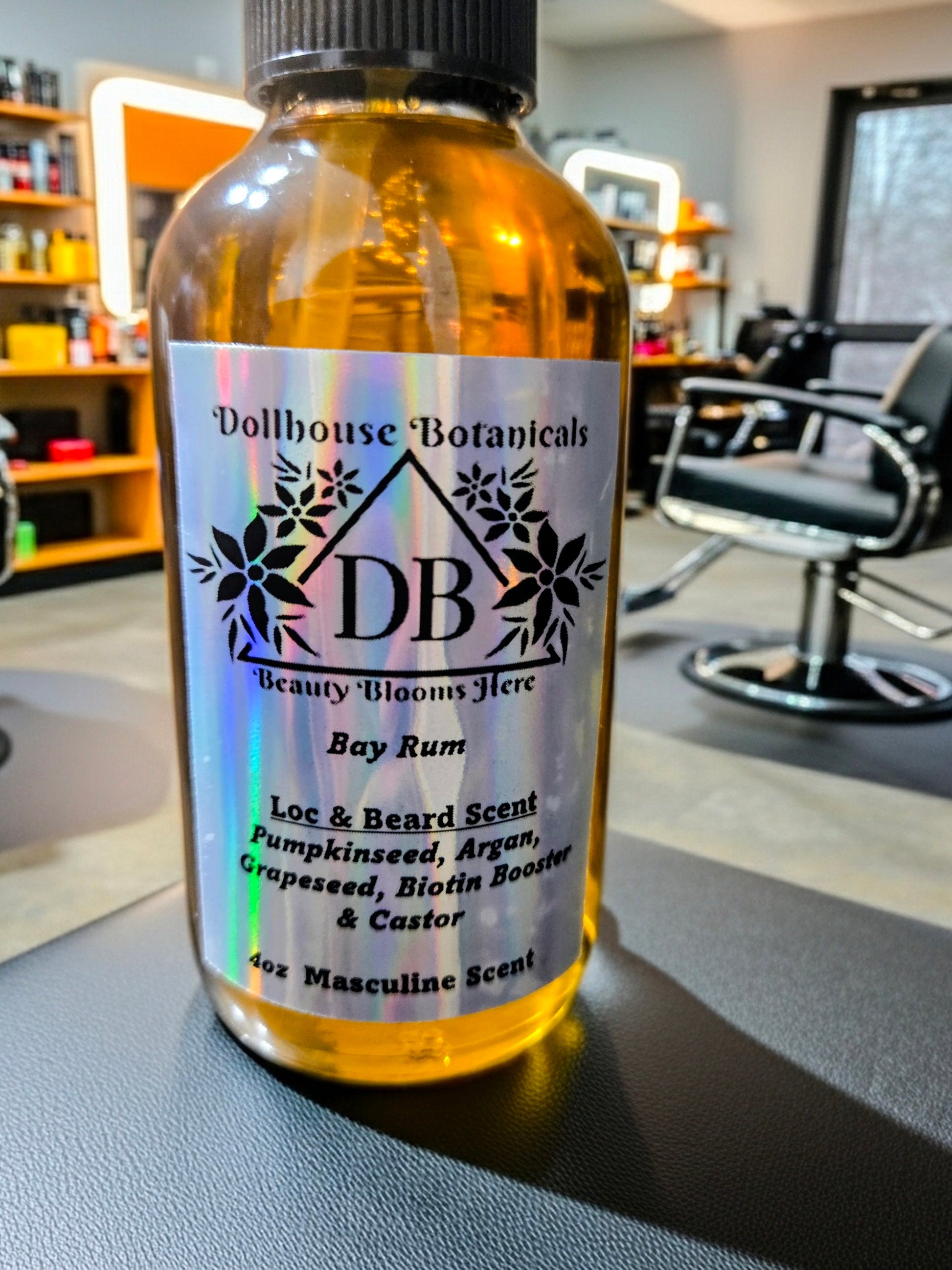 Bottle of Dollhouse Botanicals Bay Rum Loc and Beard Scent. Golden oil in glass bottle. Front facing with label and ingredients displayed, sitting on surface in barber shop.
