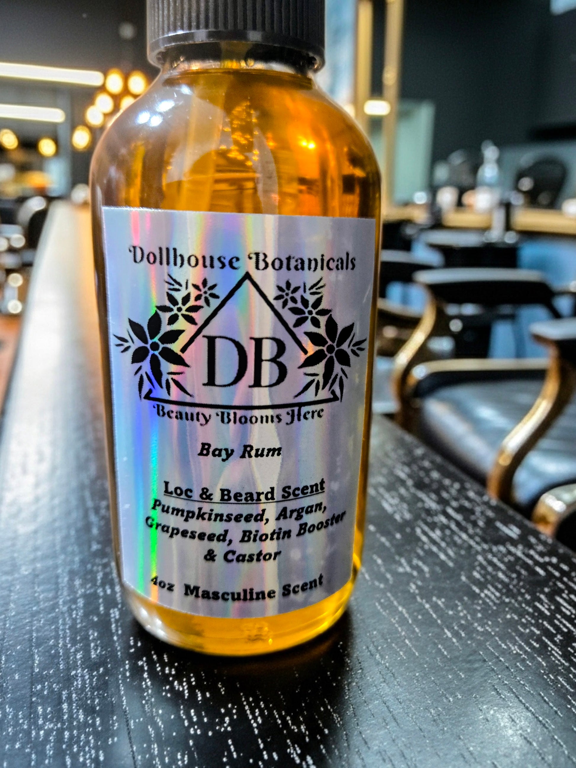 Bottle of Dollhouse Botanicals Bay Rum Loc and Beard Scent. Golden oil in glass bottle. Front facing with label and ingredients displayed, sitting on black surface in barber shop.