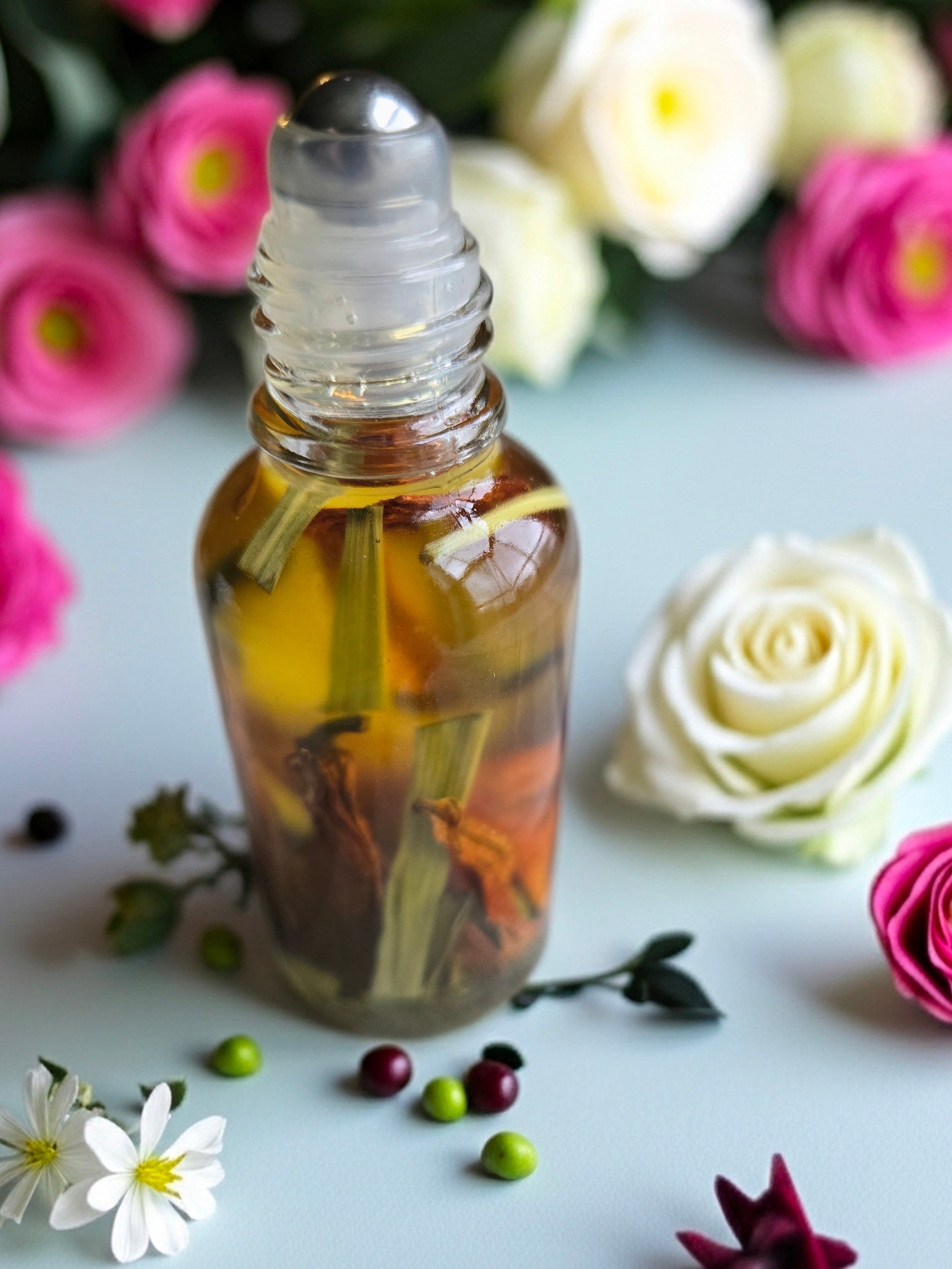 Bottle of Dollhouse Botanicals Bohemia Flora Face Hydrator. Botanicals floating in golden oil in glass bottle with steel roller ball, sitting on surface with flowers.