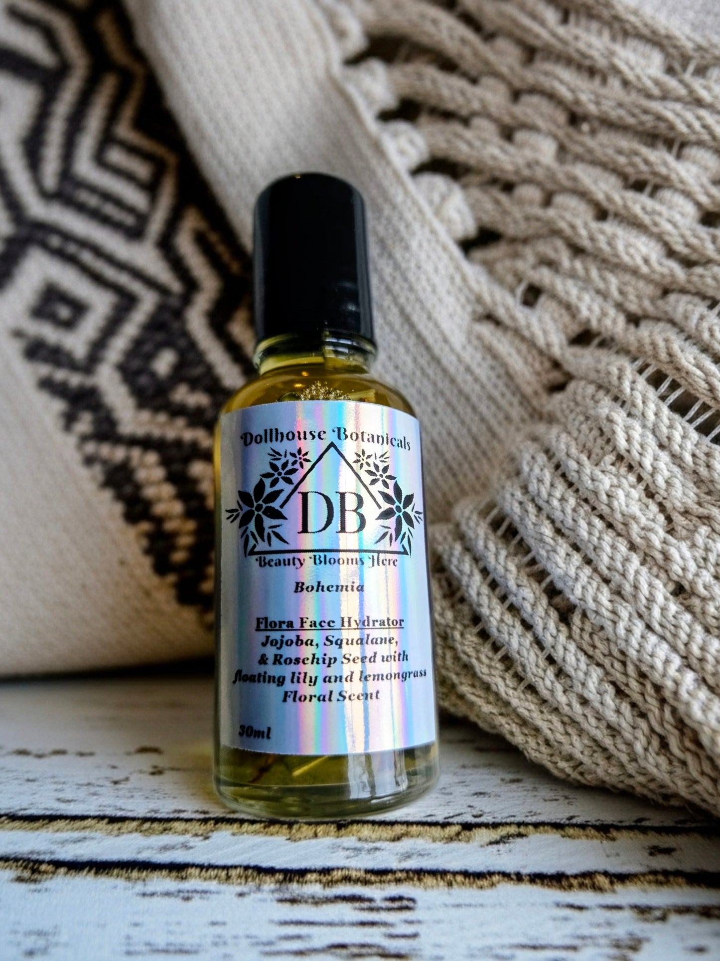 Bottle of Dollhouse Botanicals Bohemia Flora Face Hydrator. Golden oil with floating lemongrass and orange lily in glass bottle with black top. Front facing with label and ingredients displayed.