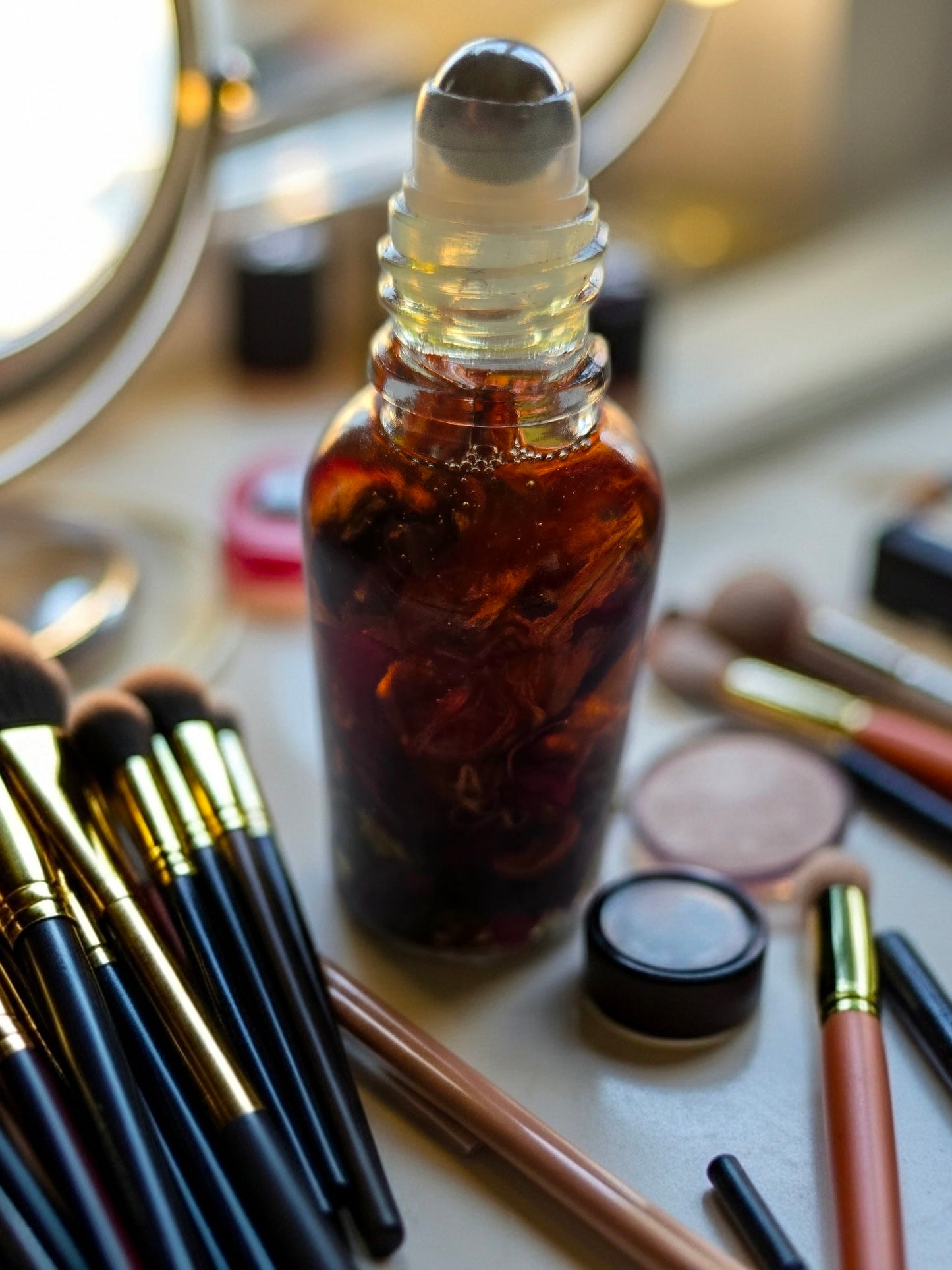 Bottle of Dollhouse Botanicals Deity Flora Face Hydrator. Golden oil with floating orange lily, purple myosotis and crushed Chines rose petals in glass bottle with steel roller ball applicator.