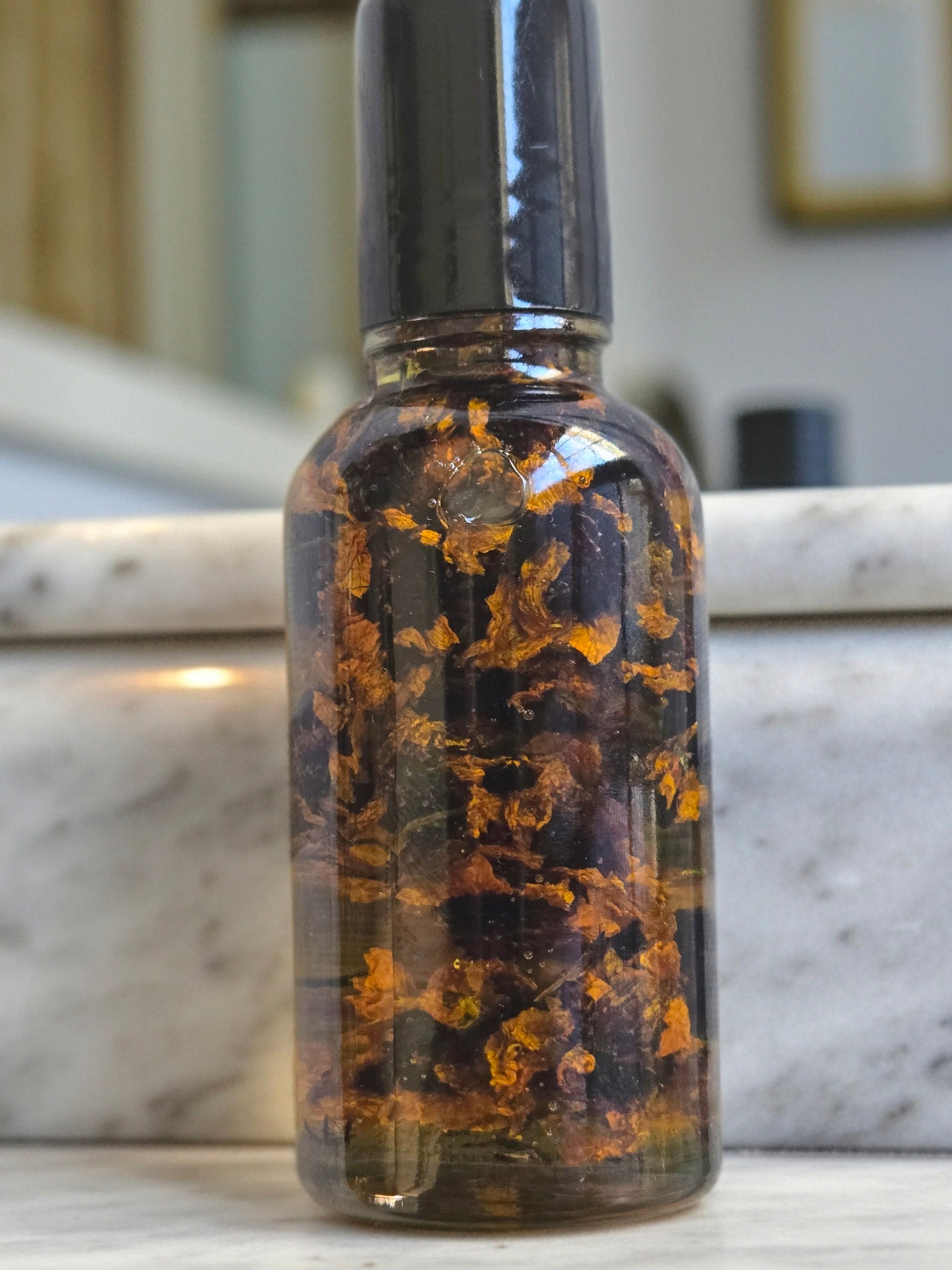 Bottle of Dollhouse Botanicals Moonlight Flora Face Hydrator. Golden oil with floating dried snow chrysanthemum in glass bottle with black top.