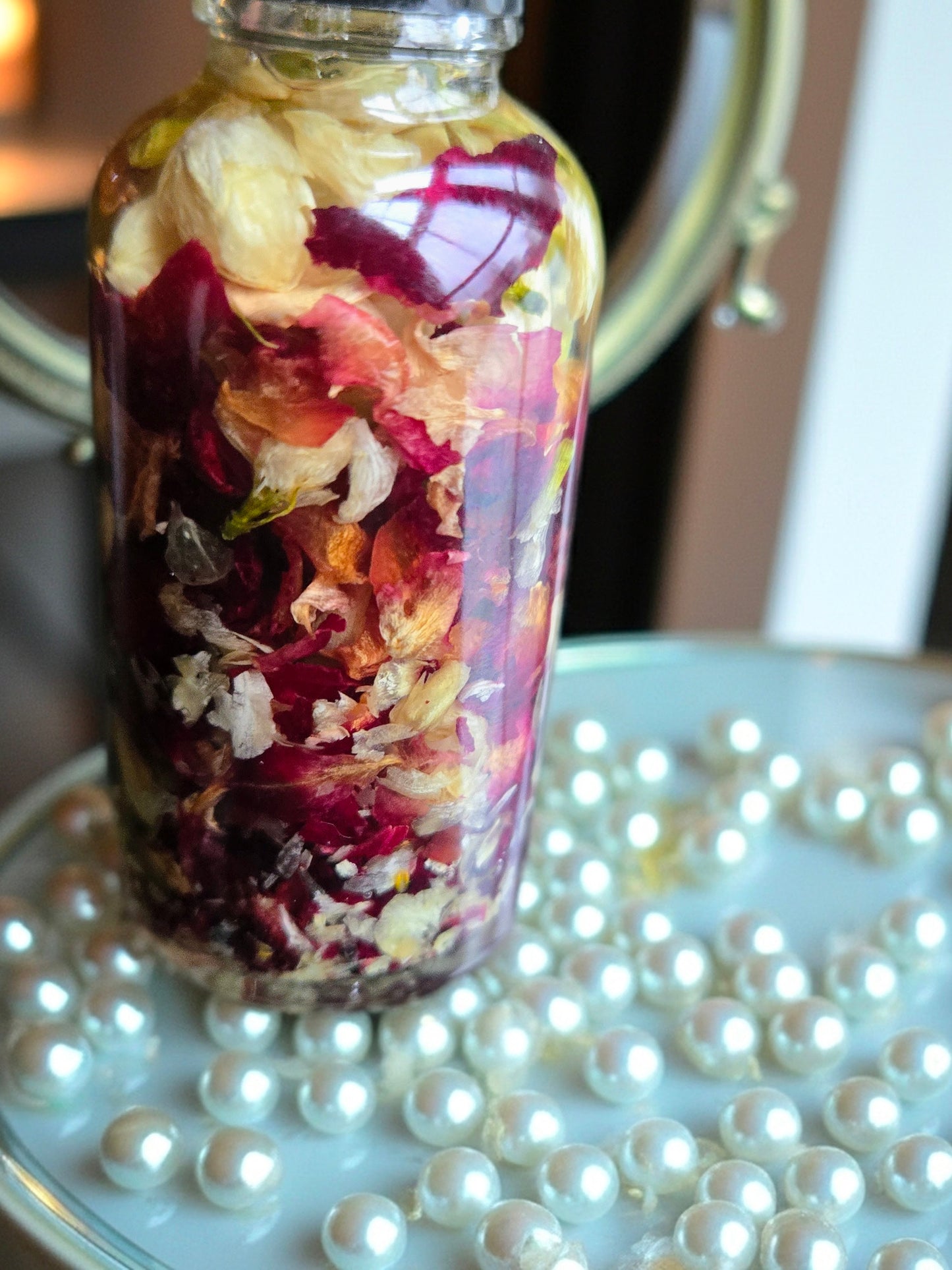 Bottle of Dollhouse Botanicals French Quarter Flora Lip Hydrator. Oil with floating dried rose petals and jasmine petals in glass bottle, sitting on surface with white pearls. Lip moisturizer.
