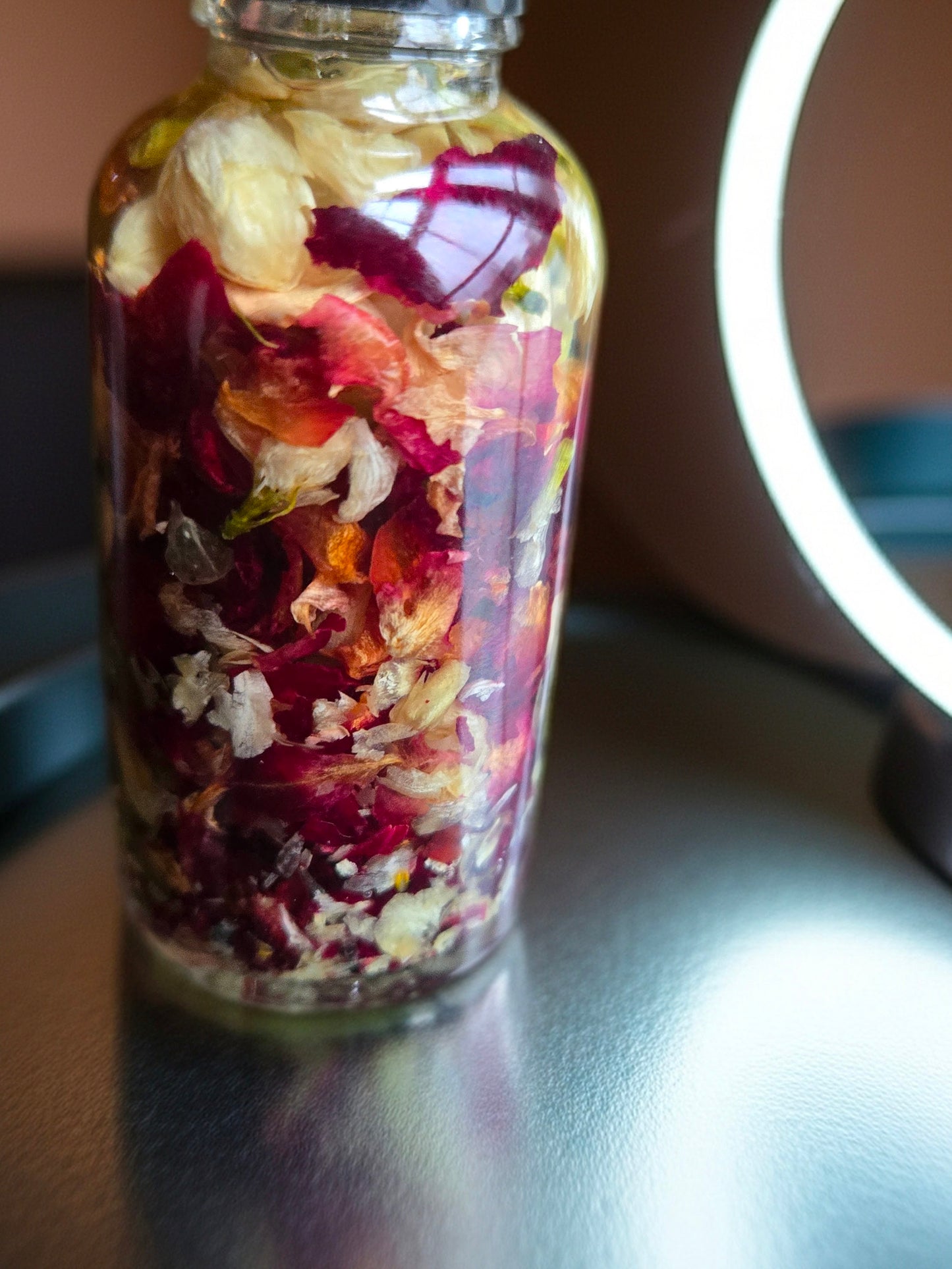 Bottle of Dollhouse Botanicals French Quarter Flora Lip Hydrator. Oil with floating dried rose petals and jasmine petals in glass bottle, sitting on vanity. Lip moisturizer.