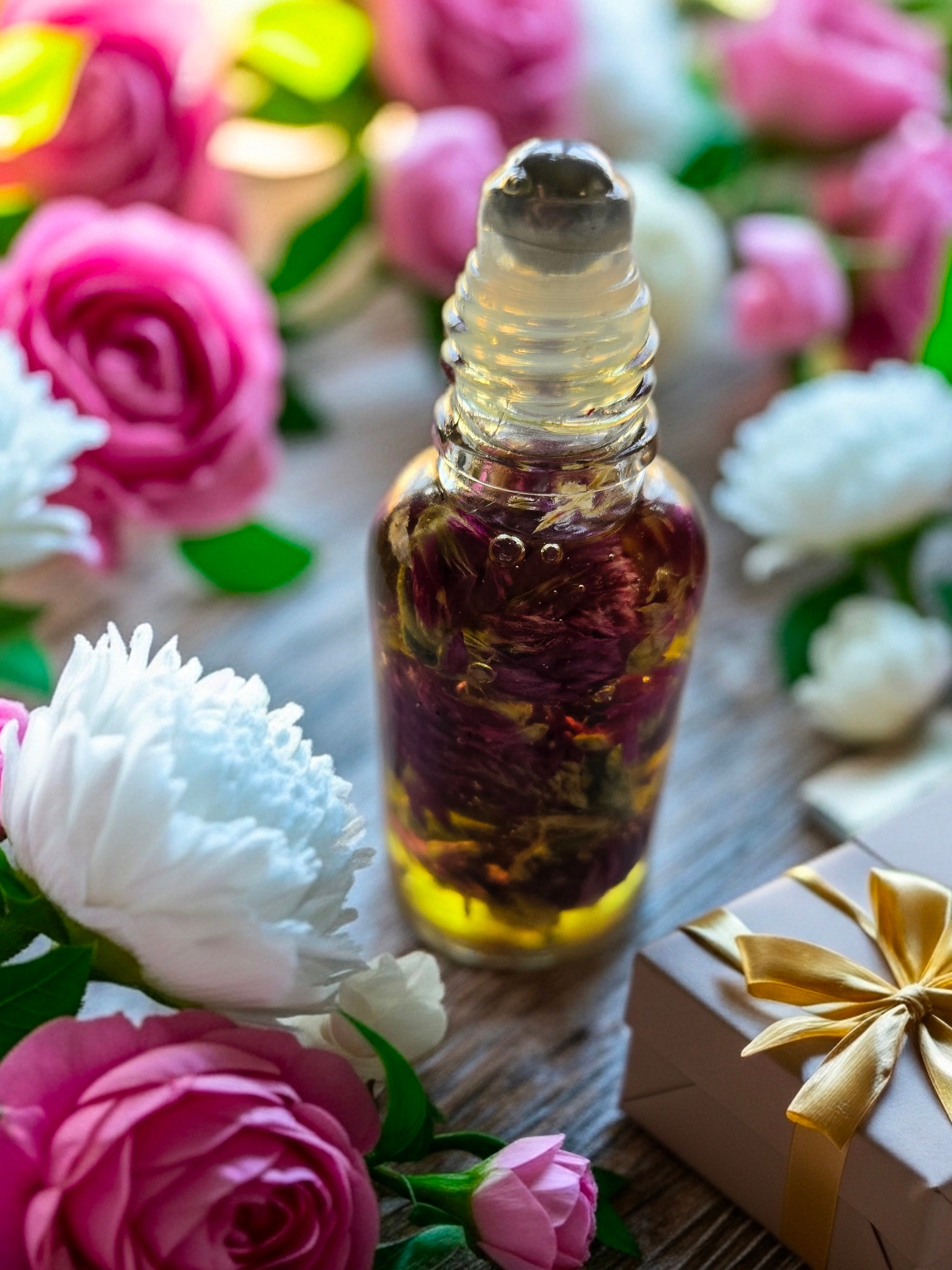 Bottle of Dollhouse Botanicals Goddess Amaranth Flora Face Hydrator. Oil with floating globe amaranth in golden oil in glass bottle with steel roll on applicator surrounded by flowers.