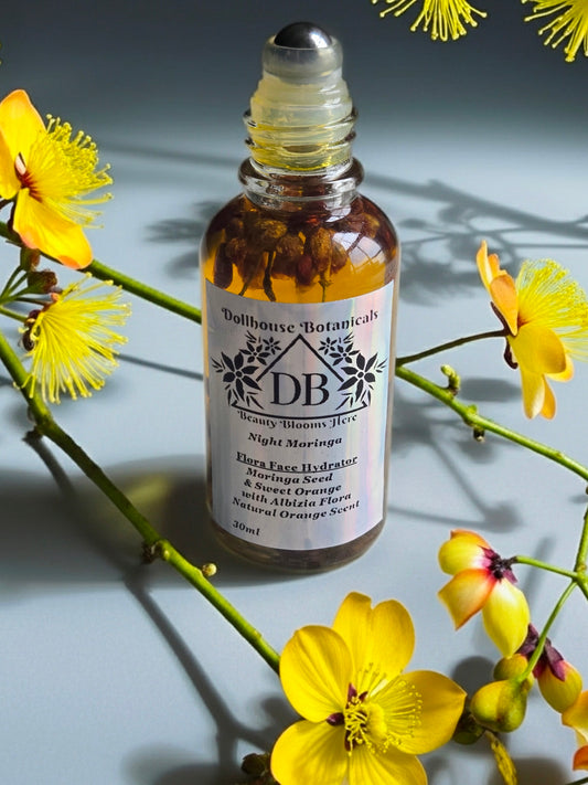 Bottle of Dollhouse Botanicals Night Moringa Flora Face Hydrator, front facing with steel roller ball applicator sitting on surface with yellow flowers.