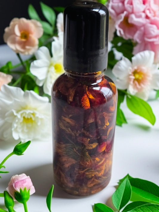 Bottle of Dollhouse Botanicals Red Sea Flora Face Hydrator. Flora floating in golden oil in glass bottle with black top, sitting on white surface with flowers.