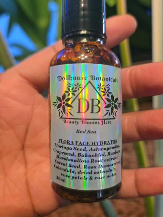 Bottle of Dollhouse Botanicals Red Sea Flora Face Hydrator. Oil with floating calendula and rose petals in glass bottle with black top lying on palm of hand.