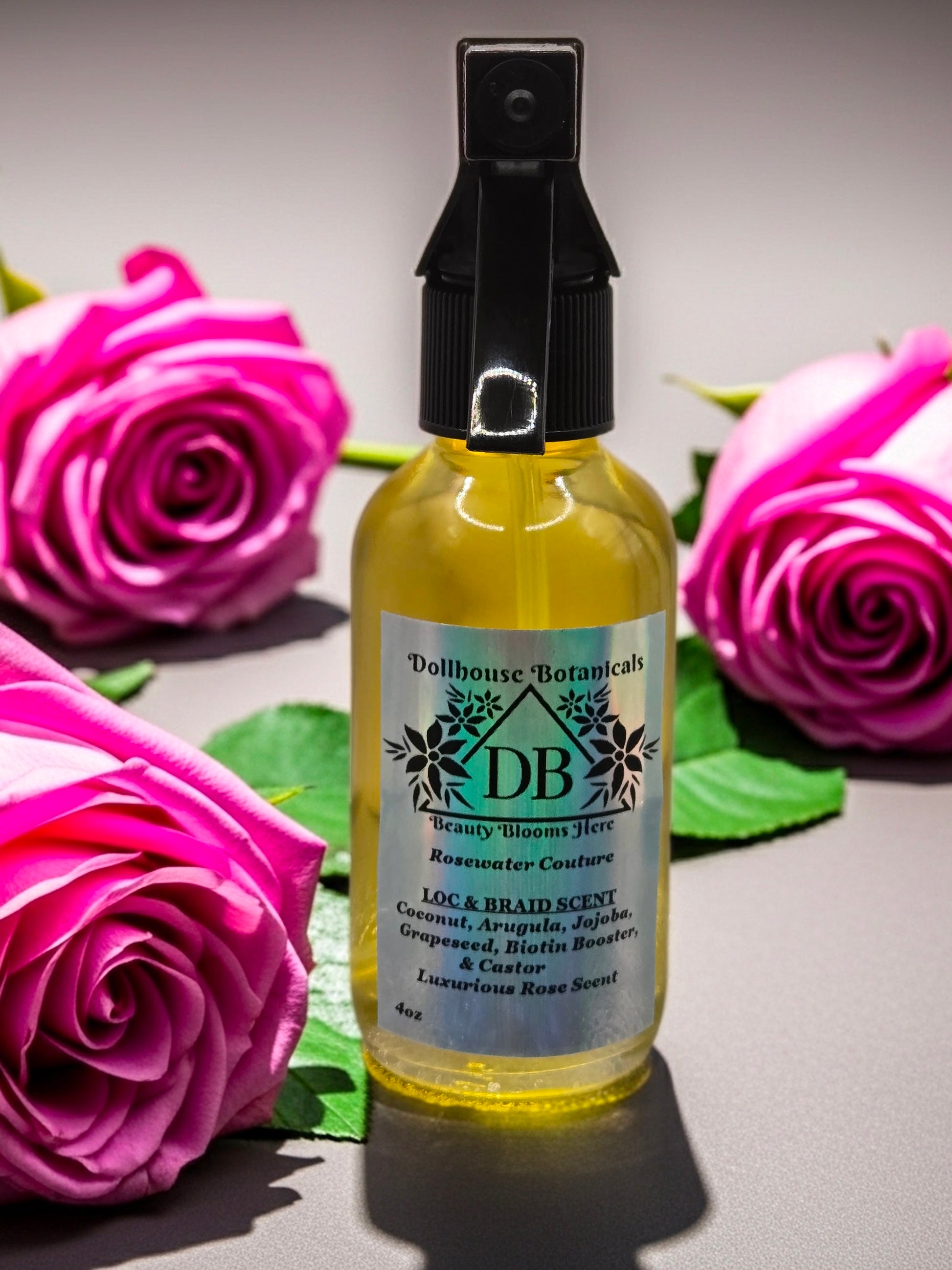 Bottle of Dollhouse Botanicals Rosewater Couture Loc and Braid Scent. Golden oil in clear glass bottle with black trigger sprayer applicator sitting on surface with pink roses.