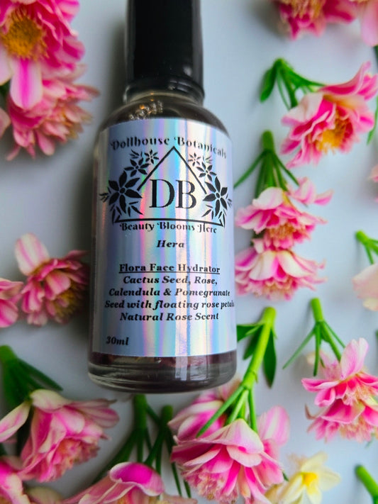 Bottle of Dollhouse Botanicals Hera Flora Face Hydrator with black top, lying front facing on surface with pink flowers.