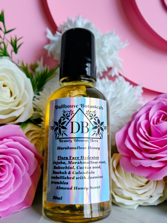 Bottle of Dollhouse Botanicals Marshmallow Honey Flora Face Hydrator with black top lying on surface with pink and white roses.