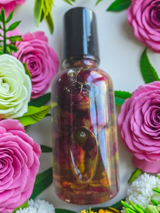 Bottle of Dollhouse Botanicals White Wine Flora Face Hydrator. Rose buds floating in golden oil in glass bottle with black top, lying on surface with roses.